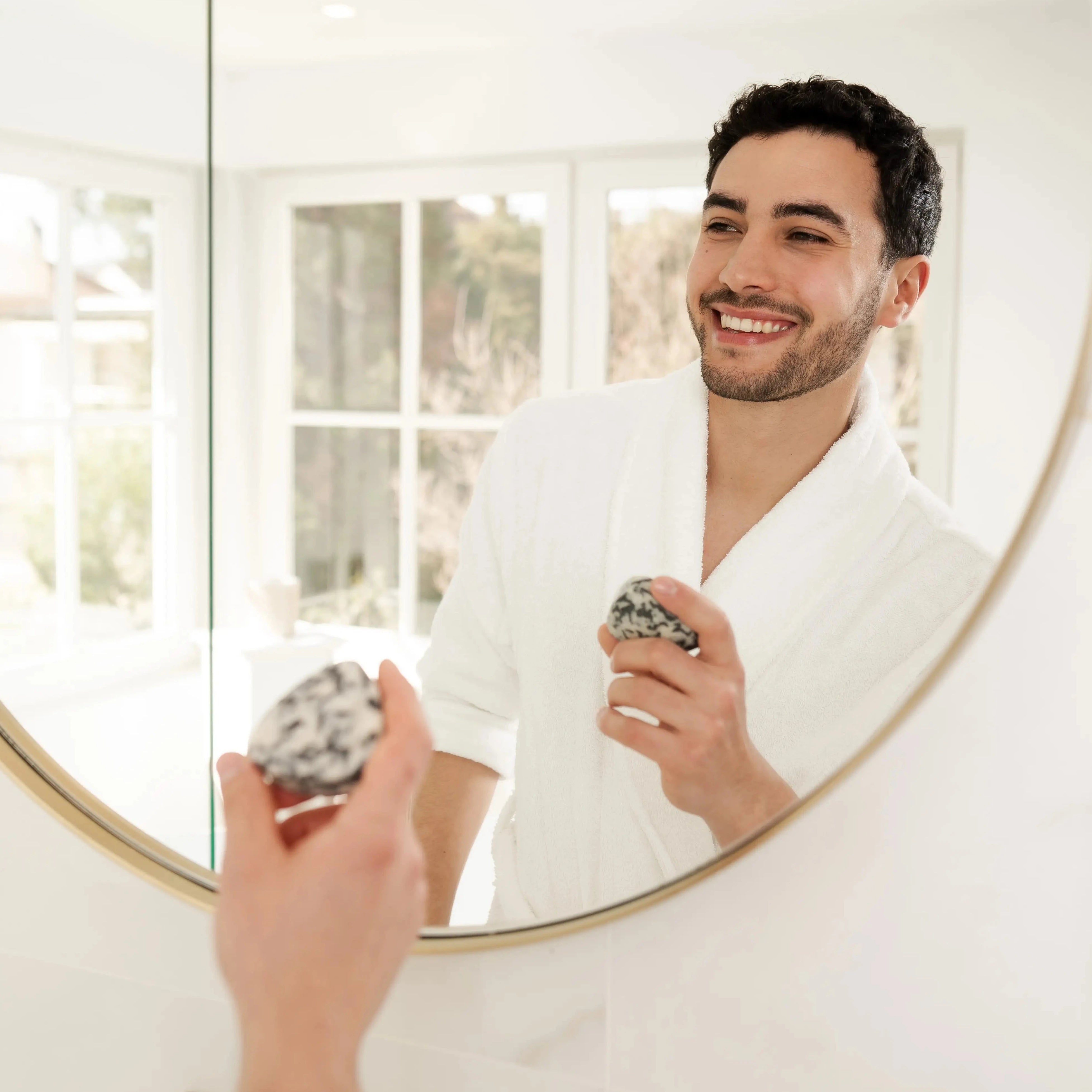 Simply Solid, männliches Model mit Solid in der Hand vor Spiegel, schwarze Haare, helle Haut, Dreitagebart, 2 in 1 Shampoo & Shower Solid, heller hintergrund, Badezimmer